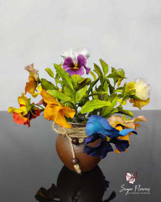 Buchet de panseluțe multicolore din zahăr, cu frunze verzi, prezentat într-un vas rustic de lut decorat cu sfoară și mărgele.